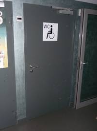 Auf dem Bild ist eine große Tür zur Behindertentoilette zu sehen. Auf der Tür ist ein weißes Schild mit der Beschriftung WC und einem Rollstuhlfahrer.