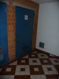 Eine große blaue Tür, umringt von einer roten Backsteinwand. Auf der Tür ist ein weißes Schild mit der Beschriftung WC und dem Symbol eines Rollstuhlfahrers. Rechts ist eine weiße Wand mit Abluftschacht zu sehen, der Boden ist mit einem rot-weißen Muster verziert.