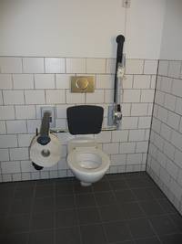 Auf dem Bild ist die Behindertentoilette im 2. Obergeschoss zu sehen. Der Toilettensitz ist weiß, hinter der Toilette ist eine schwarze Lehne. Auf beiden Seiten sind Haltebügel, der rechte ist gerade nach oben geklappt. Unter dem linken Haltebügel ist eine riesige Rolle mit Toilettenpapier. Über der Rückenlehne ist der bronzefarbene Drücker für die Spühlung. Die Wand ist mit weißen Fliesen gekachelt.
